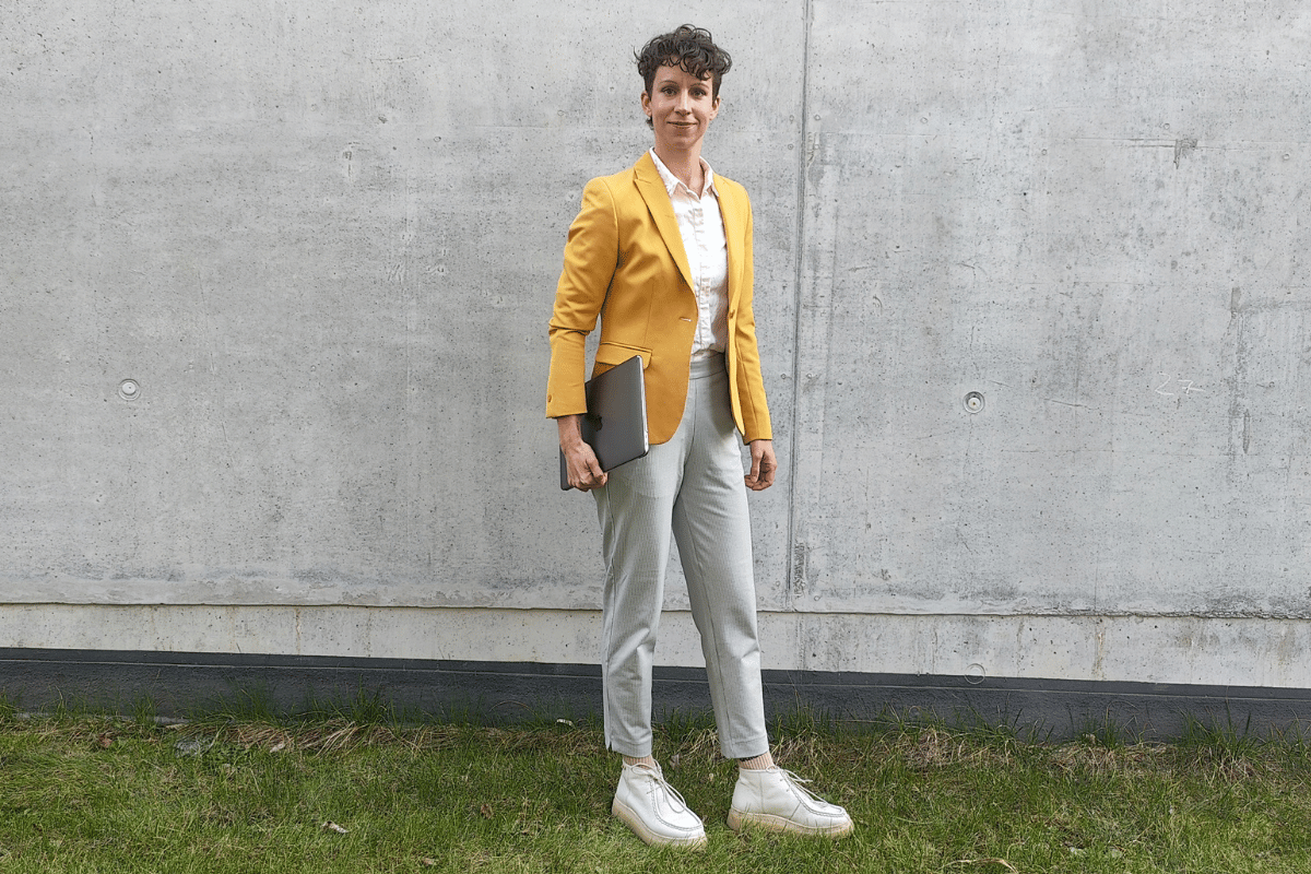 Scientist in smart dress code, holding a laptop, standing on grass in front of a raw cement wall
